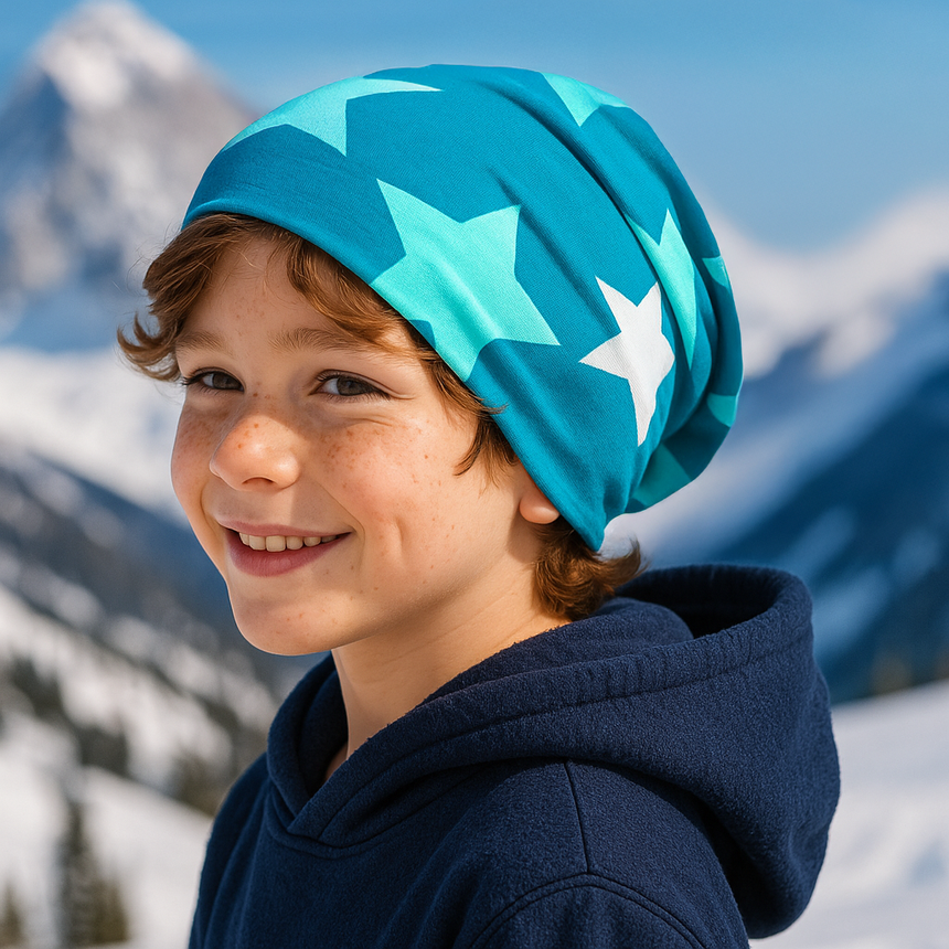 Ein Kind trägt die Wollhuhn Öko Long Beanie, zweilagig mit großen Sternen, in einer verschneiten Berglandschaft. Ideal für Komfort und Wärme im Winter.