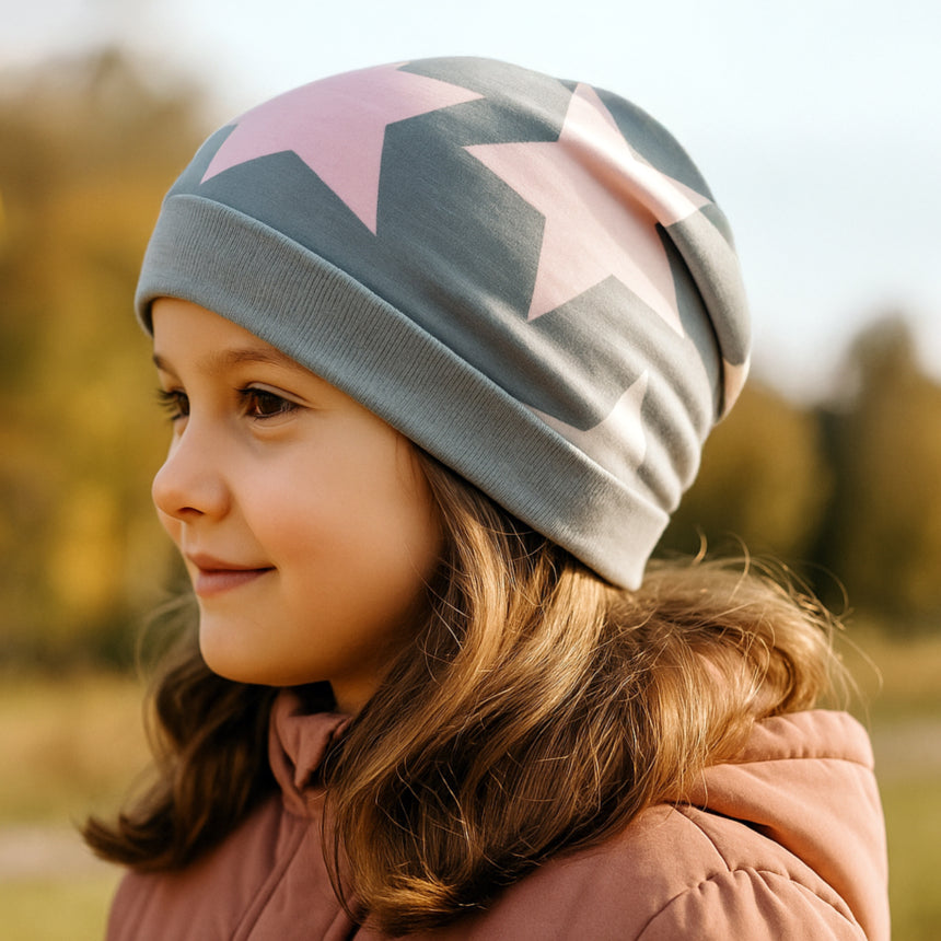 Junges Mädchen mit Wollhuhn Öko Beanie-Mütze, bedeckt mit großen pastellfarbenen Sternen, trägt staubrosafarbene Jacke in herbstlicher Umgebung, strahlt Ruhe aus.