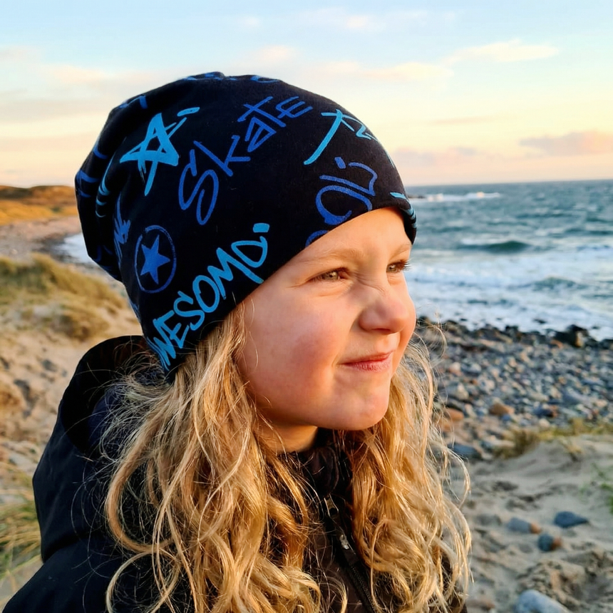 Kind am Strand trägt Wollhuhn Öko Long Beanie, Wendemütze mit Graffiti-Motiven. Perfekt für Übergangszeiten, handgemacht in Deutschland aus weichem Öko-Jersey.