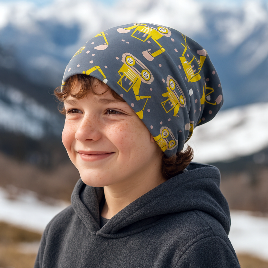 Junge trägt Wollhuhn Öko Long Beanie mit gelben Baggermotiven, ideal für ganzjähriges Tragen, zweilagig und wendbar, in einer verschneiten Berglandschaft.