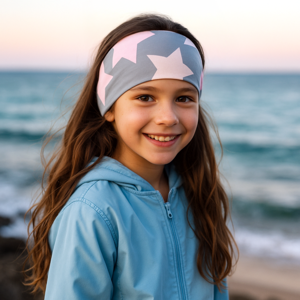 Mädchen am Strand trägt Wollhuhn Elastisches Öko Stirnband/Haarband Jersey Zweilagig Breit Big Stars Grau/Rosa, lächelt in die Kamera, Meer im Hintergrund.