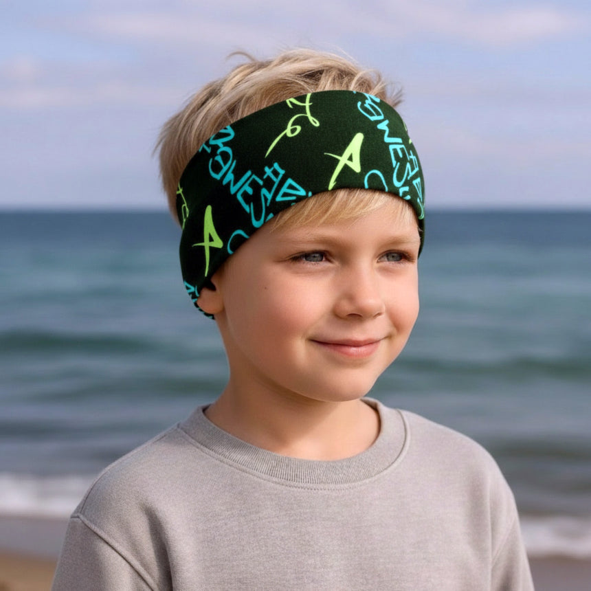 Junger Junge am Meer trägt das Wollhuhn Elastisches Öko Stirnband in Schwarz/Blau/Grün, ideal für Outdoor-Abenteuer und sportliche Aktivitäten.