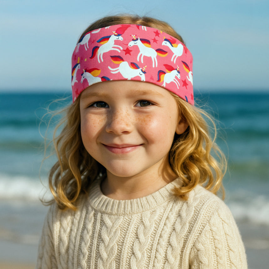 Mädchen am Strand trägt Wollhuhn Elastisches Öko Stirnband Happy Unicorn, handgemacht aus weichem, dehnbaren Jersey, ideal für Outdoor-Aktivitäten.