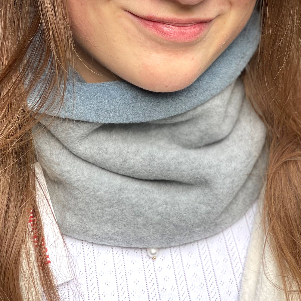 Nahaufnahme einer Frau, die den Wollhuhn WINTER Halstuch/Schlupfschal „Unser Wärmster“ aus zwei Lagen Fleece in Hellgrau/Rauchblau trägt.