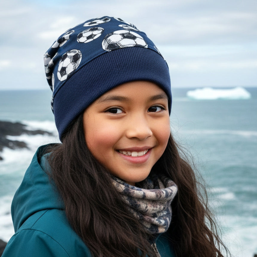 Junges Mädchen trägt Wollhuhn Dünne Öko Beanie-Mütze Sommer/Übergang Einlagig Fußball Dunkelblau am Meer, ideal für kühle Tage und sportliche Aktivitäten.