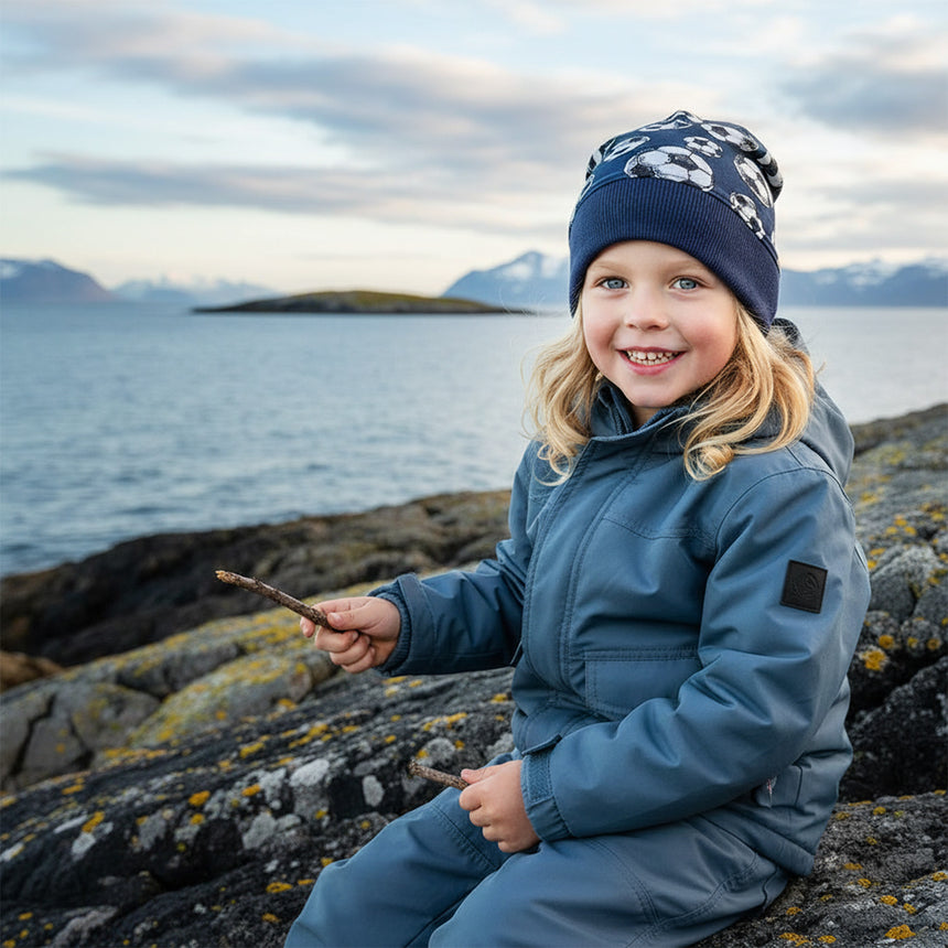 Kind am ruhigen Ufer, trägt die Wollhuhn Dünne Öko Beanie-Mütze Sommer/Übergang Einlagig Fußball Dunkelblau. Lächelt, hält Stöcke. Hintergrund: Berge, Wasser.