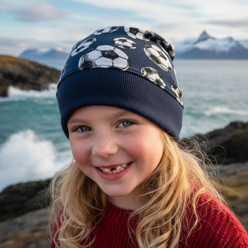 Junges Mädchen trägt Wollhuhn Dünne Öko Beanie-Mütze Sommer/Übergang Einlagig Fußball Dunkelblau an felsiger Küste, lächelt in die Kamera.