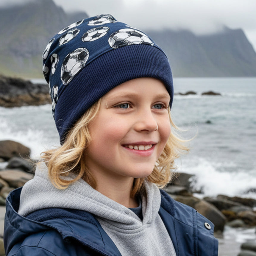 Ein Kind am felsigen Ufer trägt eine Wollhuhn Dünne Öko Beanie-Mütze Sommer/Übergang Einlagig Fußball Dunkelblau, perfekt für kühle Tage und sportliche Aktivitäten.