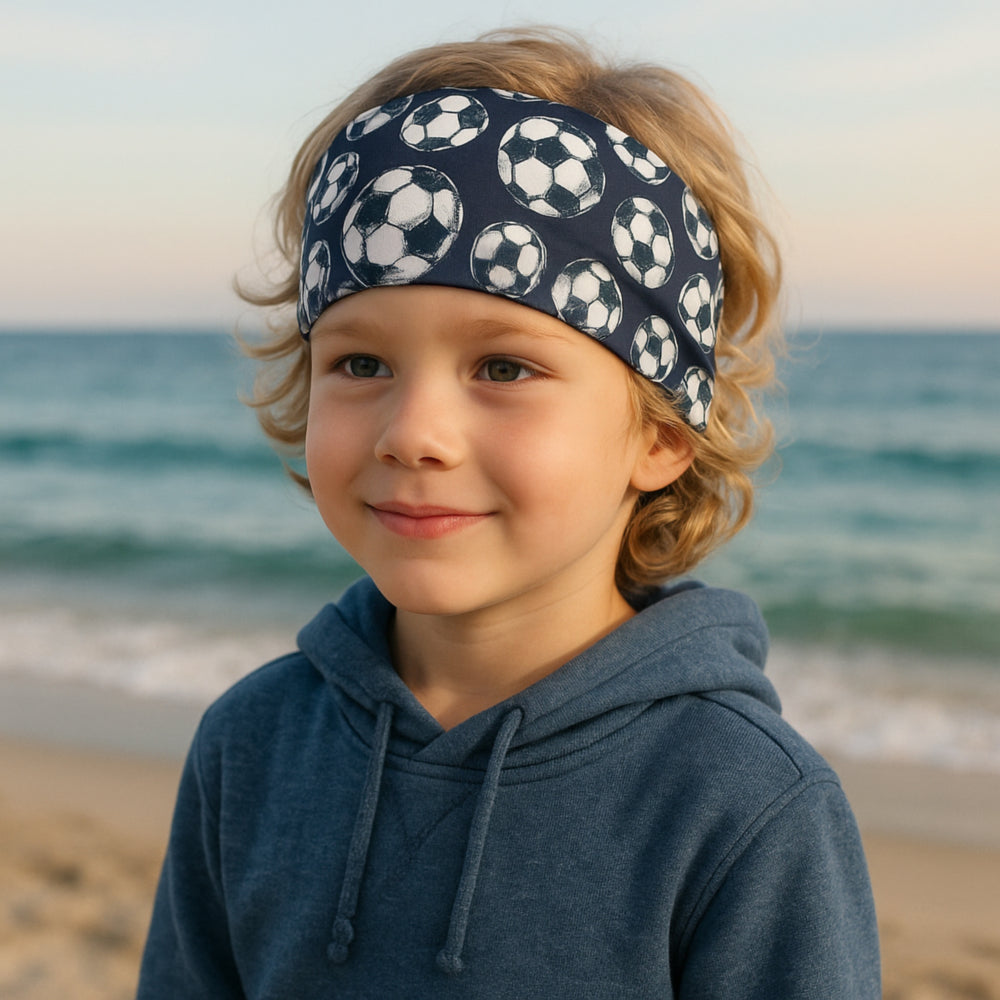 Kind am Strand trägt das Wollhuhn Elastisches Öko Stirnband, breit und zweilagig, mit Fußballmotiv. Handgemacht, bequem und vielseitig für Outdoor-Aktivitäten.
