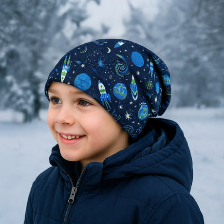 Kind mit Wollhuhn Öko Long Beanie in Space/Rakete-Design, lächelt im verschneiten Wald. Handgemachte, wendbare Mütze, ideal für milde Wintertage, bequem und elastisch.