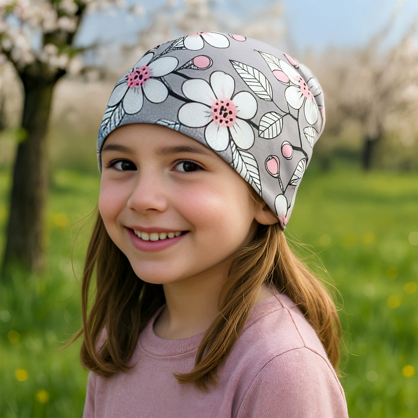 Mädchen trägt Wollhuhn Öko Shorties Wendemütze, ganzjährig und zweilagig, im Stil Kirschblüten Grau/Rosa. Sie lächelt und zeigt die bequeme, mitwachsende Passform.