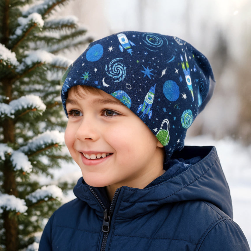 Wollhuhn Öko Shorties: Kind trägt blaue, zweilagige Wendemütze vor schneebedecktem Baum. Handgemacht, bequem, ideal für aktive Kinder, passt unter Helme.