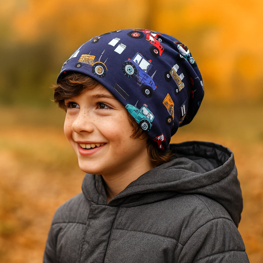 Ein Kind trägt die Wollhuhn Öko Long Beanie, eine wendbare, zweilagige Mütze mit bunten Fahrzeugmotiven, ideal für ganzjähriges, komfortables Tragen.