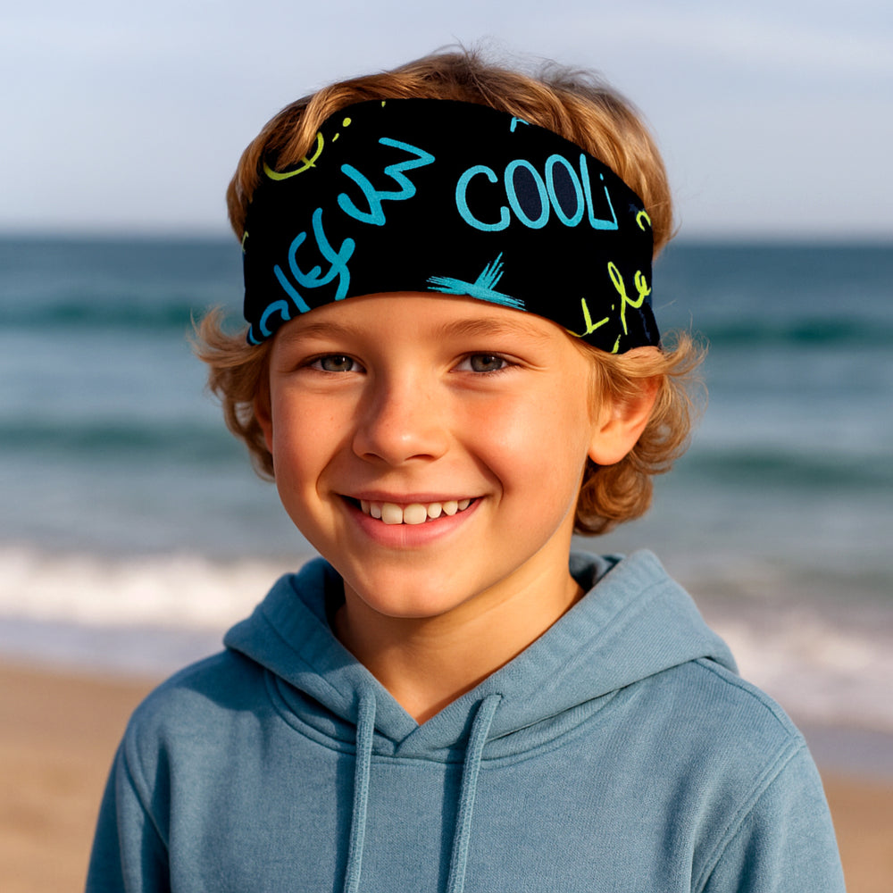 Junge am Strand trägt das Wollhuhn Elastisches Öko Stirnband - handgemachtes, zweilagiges Jersey-Stirnband, ideal für Outdoor-Aktivitäten, bequem und vielseitig tragbar.