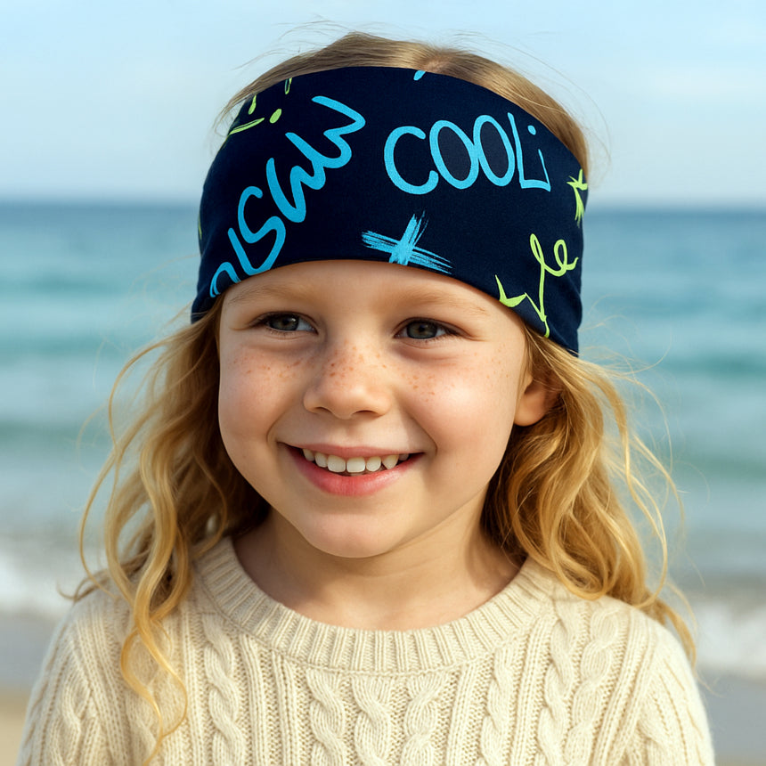 Mädchen am Strand trägt Wollhuhn Elastisches Öko Stirnband, Jersey zweilagig, ideal für Outdoor-Aktivitäten. Komfortabel und handgemacht aus Bio-Baumwolle.
