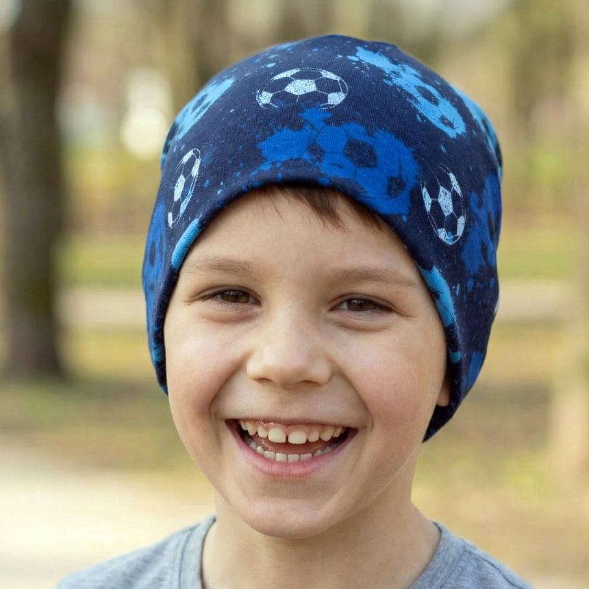 Junger Junge im Park, trägt Wollhuhn Öko Shorties Wendemütze mit Fußballmotiv; handgemacht, zweilagig, ideal für aktive Kinder, hält Ohren warm.
