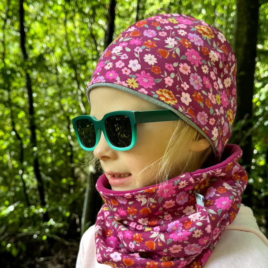 Mädchen im Wald trägt Wollhuhn Öko Shorties, eine zweilagige, wendbare Beanie mit floralem Muster, passend für jede Jahreszeit.