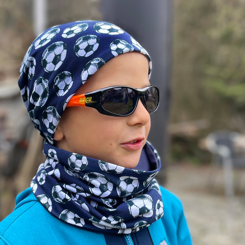 Kind mit Wollhuhn Öko Shorties, zweilagiger Wendemütze in Fußballprint, trägt schwarze Sonnenbrille. Perfekt für aktive Kinder, handgemacht in Deutschland.