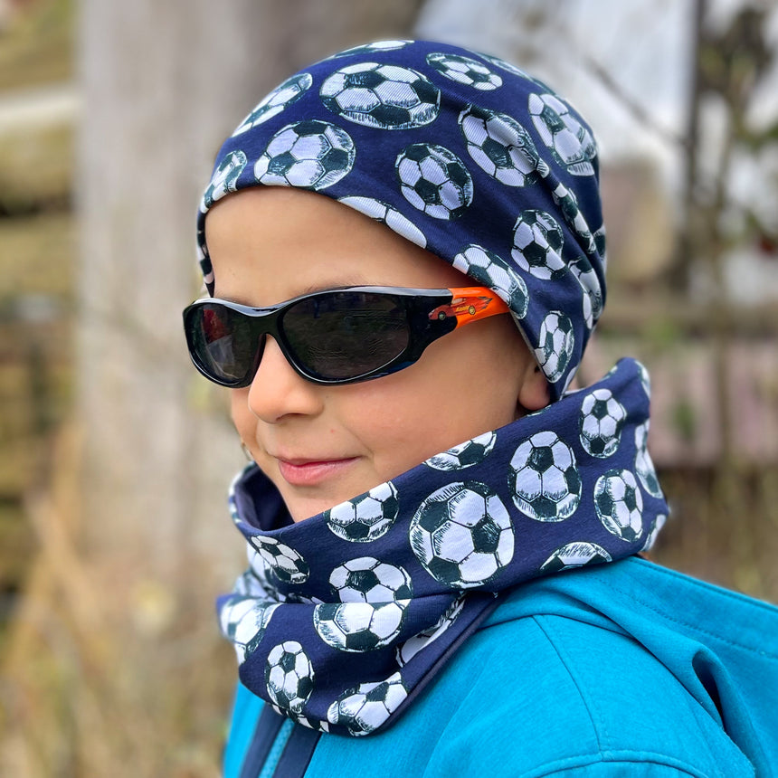 Ein Kind trägt die Wollhuhn Öko Shorties Wendemütze mit Fußballmuster und passendem Halswärmer, ergänzt durch eine blaue Jacke und schwarze Sonnenbrille.