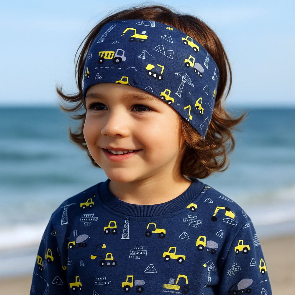 Ein Kind am Strand trägt das Wollhuhn Elastisches Öko Stirnband, Jersey zweilagig, mit Baustellen- und Baggermotiv, dunkelblau/gelb, bequem und handgemacht.