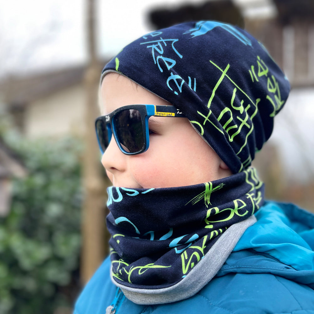 Junge trägt Wollhuhn Öko Shorties, eine zweilagige Wendemütze in Schwarz, kombiniert mit Sonnenbrille und Schal. Perfekt für aktive Kinder, handgefertigt in Deutschland.