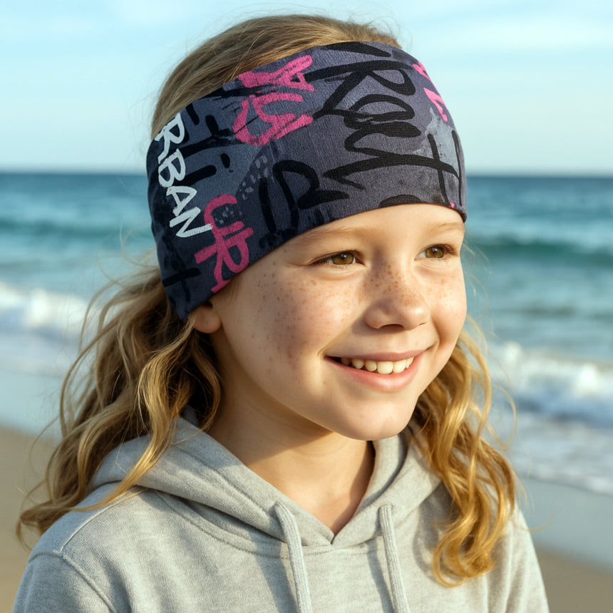 Mädchen am Strand trägt Wollhuhn Elastisches Öko Stirnband aus Jersey, zweilagig, breit, ideal für Outdoor-Aktivitäten, handgemacht in Deutschland aus Bio-Baumwolle.