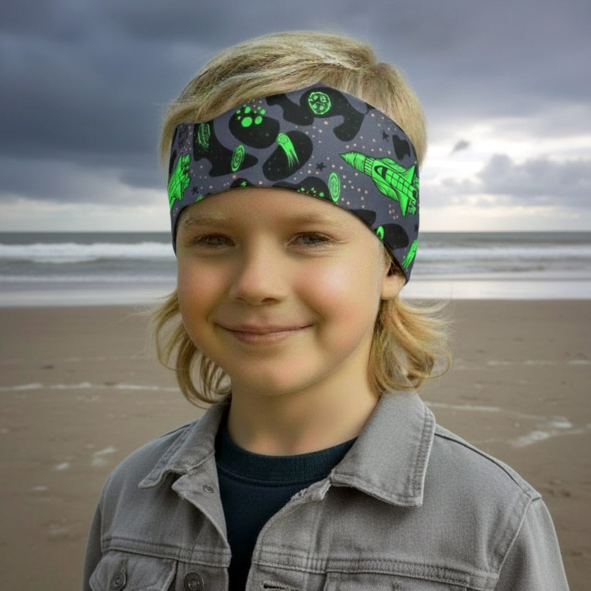 Kind am Strand mit Wollhuhn Elastisches Öko Stirnband Jersey Zweilagig Breit Space/Rakete Neon Grün, lächelt, trägt dunkles Shirt und graue Jacke.