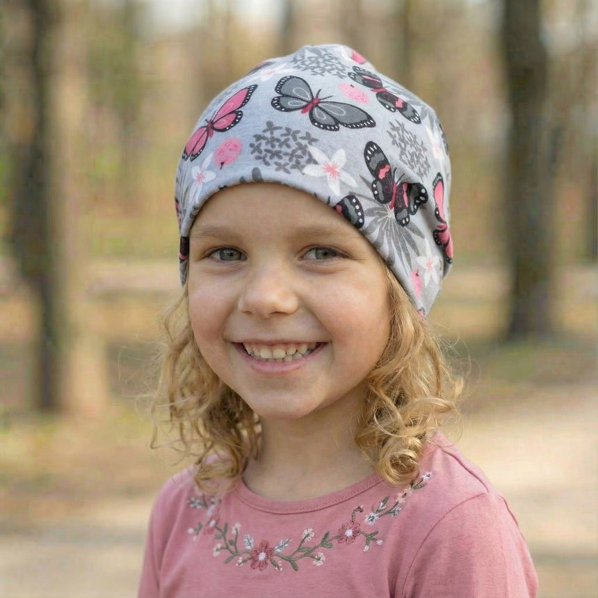 Mädchen trägt Wollhuhn Öko Shorties/Wendemütze, zweilagig mit Schmetterlingen, in einem Wald. Handgemacht, bequem und für aktive Kinder geeignet.