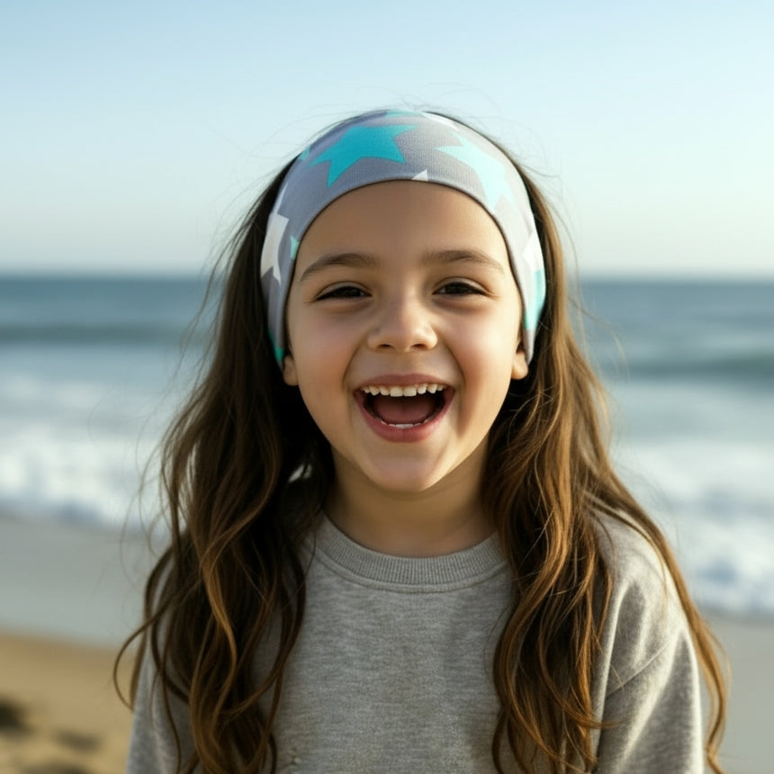 Mädchen am Strand mit Wollhuhn Elastisches Öko Stirnband/Haarband Jersey Zweilagig Breit Big Stars Sterne Grau/Mint, lächelnd, mit Meereshintergrund.