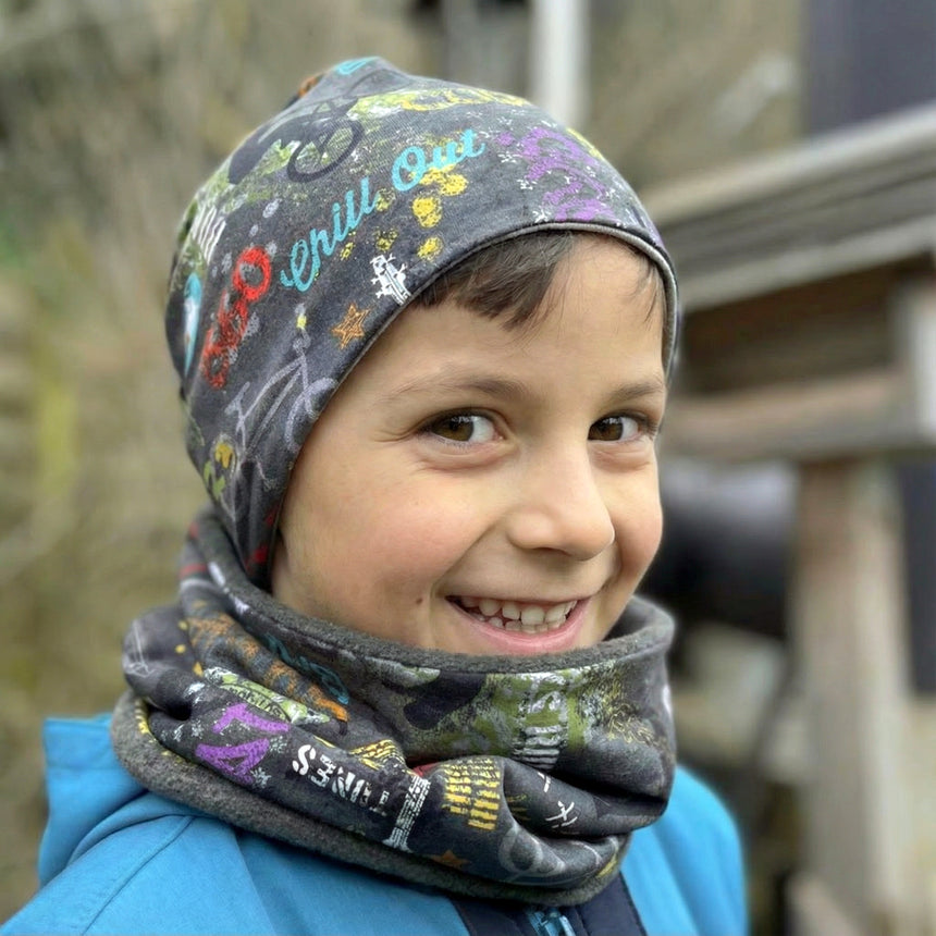 Junge trägt Wollhuhn Öko Shorties, Wendemütze, Cool Stuff Anthrazit, lächelnd im Freien, mit grauem Beanie und Halswärmer, passend unter Helme, handgemacht in Deutschland.