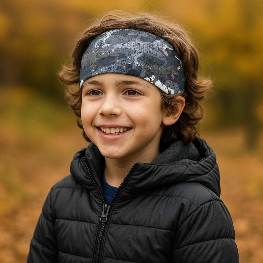 Junge mit Wollhuhn Elastisches Öko Stirnband, das die Haare zurückhält. Er trägt eine dunkel gesteppte Jacke, lächelt und steht in einer herbstlichen Umgebung.