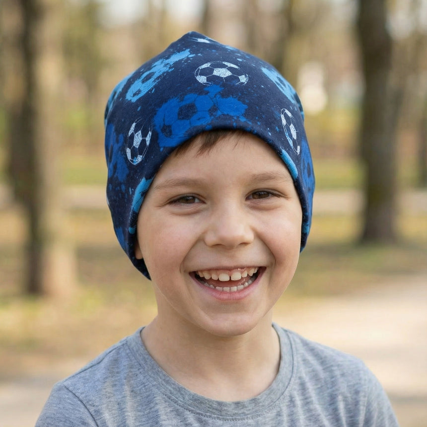Junge mit Wollhuhn Öko Shorties Wendemütze, Fußball Blau, lächelt im Freien. Handgemachte, zweilagige Jersey-Beanie, bequem und ideal für aktive Kinder.
