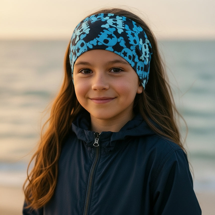 Junges Mädchen am Strand trägt Wollhuhn Elastisches Öko Stirnband / Haarband Jersey Zweilagig Breit Coole Skulls Blau/Schwarz, perfekt für Outdoor-Aktivitäten.