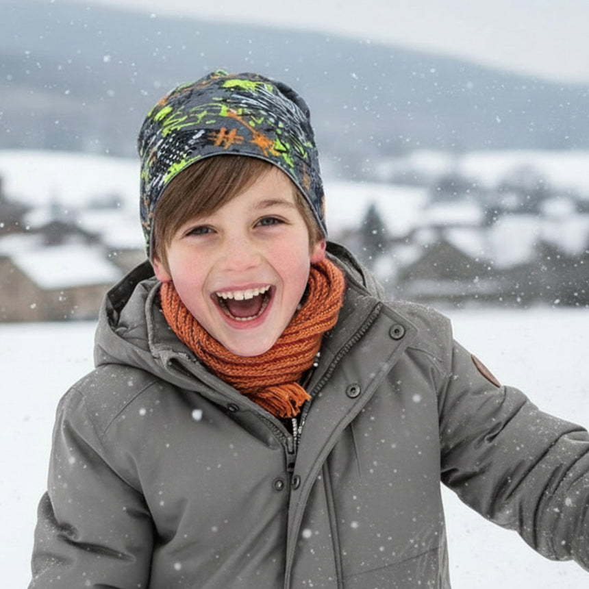 Kind mit Wollhuhn Öko Long Beanie Wendemütze, Graffiti Grau/Bunt. Lächelnd im Schnee, trägt eine graue Jacke, orange Schal, und bunte Mütze.