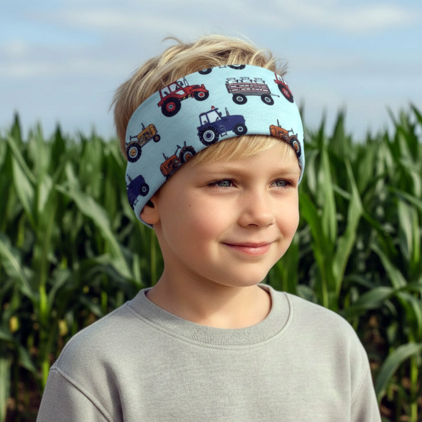 Junger Junge mit einem Wollhuhn Elastisches Öko Stirnband, zweilagig breit, mit Traktor-Motiven, stehend vor einem grünen Maisfeld, lächelnd.