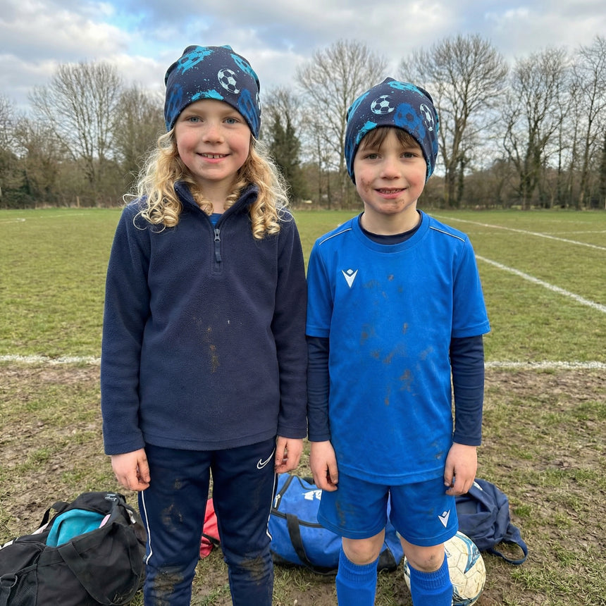 Zwei Kinder tragen Wollhuhn Öko Long Beanie mit Fußballmotiv auf einem schlammigen Sportfeld, perfekt für Komfort und Wärme bei Outdoor-Aktivitäten, handgefertigt in Deutschland.