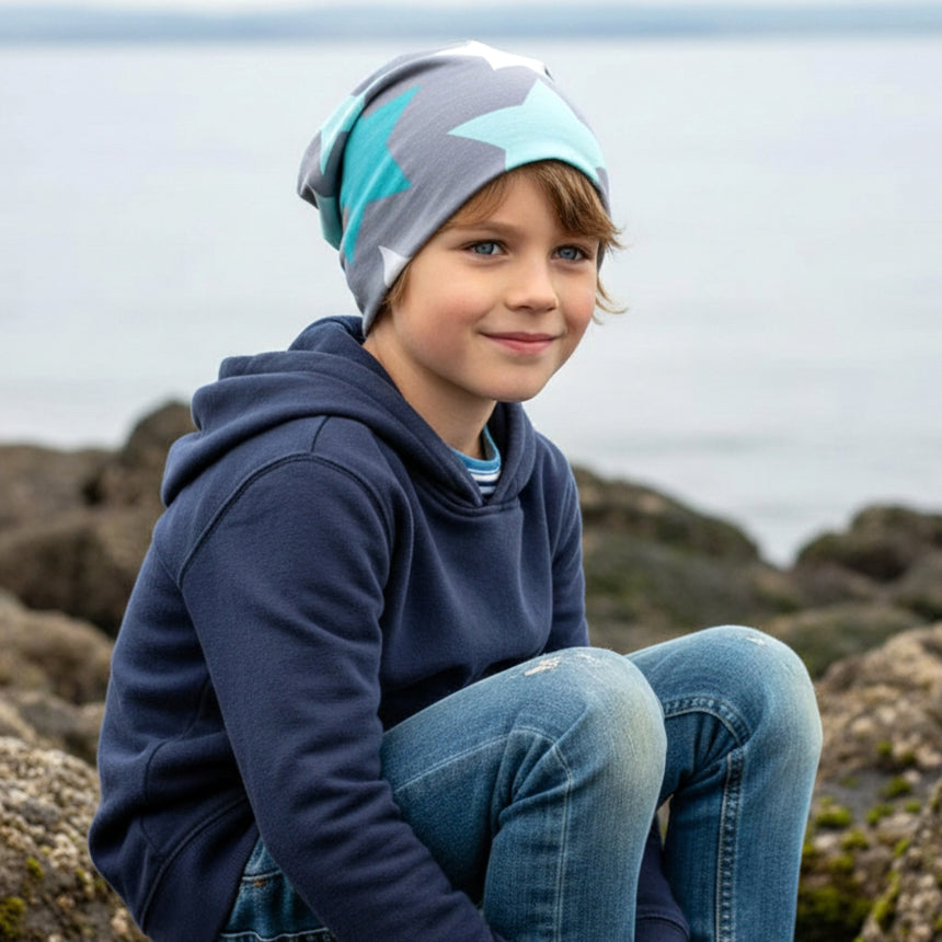 Junge trägt Wollhuhn Öko Long Beanie mit großen grauen und mintfarbenen Sternen, sitzt entspannt auf Küstenfelsen, genießt ruhigen Moment am Meer.