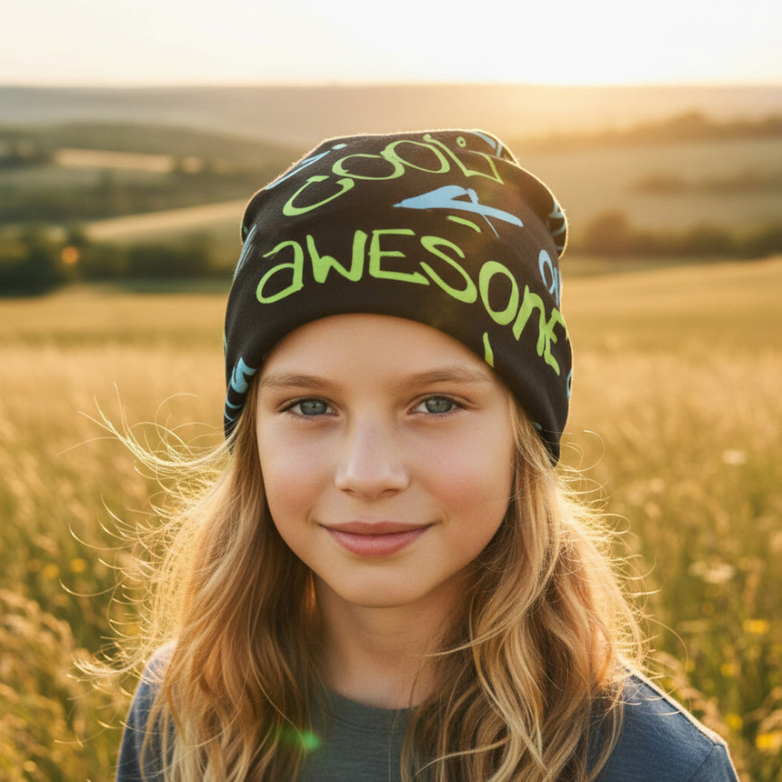 Ein Mädchen trägt die Wollhuhn Öko Wendemütze mit awesome-Schriftzug, ideal für ganzjähriges Tragen, aus weichem Baumwolljersey, in einem sonnigen Feld.