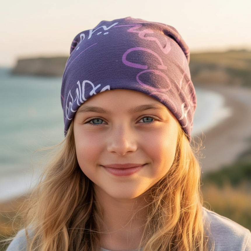 Mädchen lächelt in Kamera, trägt Wollhuhn Öko Long Beanie / Wendemütze in Violett, vor Küste. Handgemachte, zweilagige Mütze, ideal für Übergangszeiten.