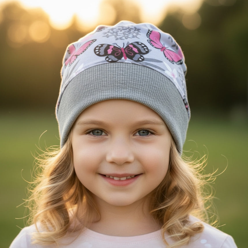 Ein lächelndes Mädchen trägt die Wollhuhn Dünne Öko Beanie-Mütze Sommer/Übergang mit Schmetterlingsmuster, handgemacht aus weichem, elastischem Öko-Jersey für Komfort und Stil.