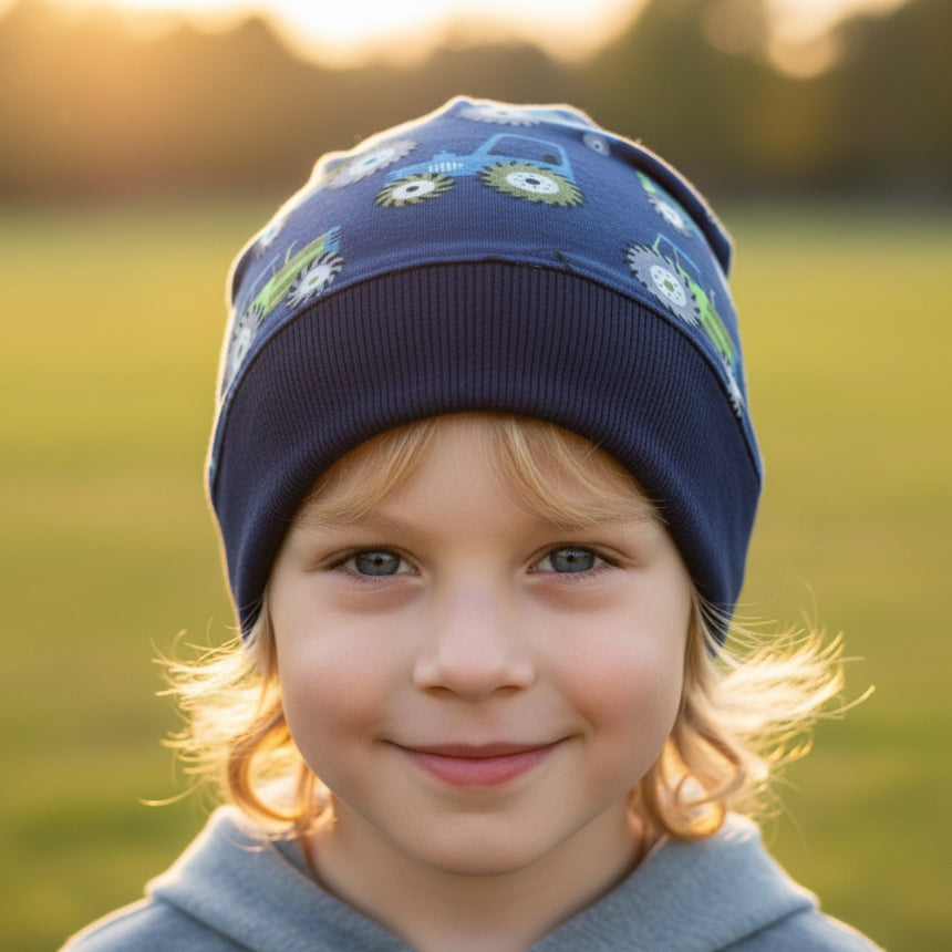 Ein lächelndes Kleinkind trägt die Wollhuhn Dünne Öko Beanie-Mütze Sommer/Übergang Einlagig Traktor/Trecker Dunkelblau auf einer Wiese im goldenen Abendlicht.