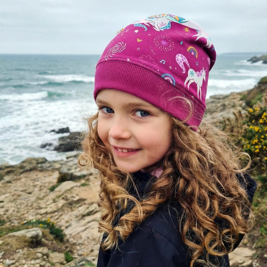 Junges Kind am felsigen Meeresufer, trägt Wollhuhn Dünne Öko Beanie-Mütze Sommer/Übergang Einlagig Einhorn/Einhörner Beere/Pink, strahlt mit einem schüchternen Lächeln.