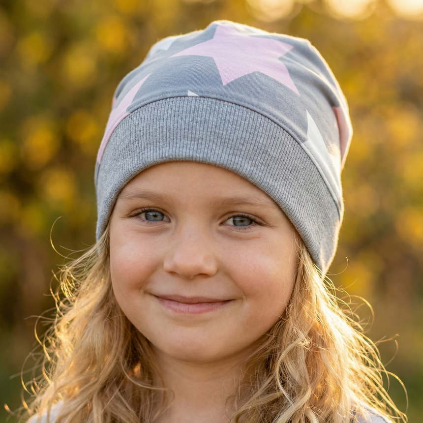 Mädchen mit blonden Haaren trägt Wollhuhn Dünne Öko Beanie-Mütze Sommer/Übergang Einlagig Big Stars Sterne Grau/Rosa, lächelt draußen in warmem Licht.
