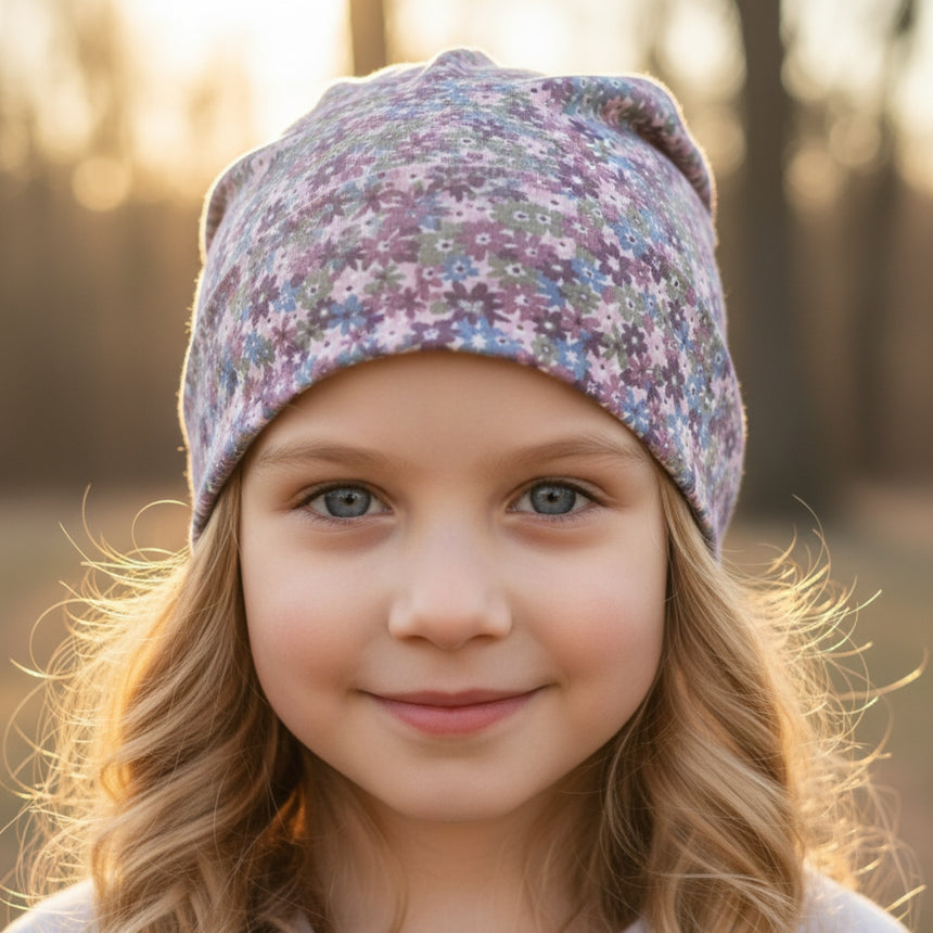 Ein junges Mädchen trägt die Wollhuhn Öko Long Beanie, Wendemütze, Ganzjährig, Zweilagig, Blümchen Dark Pastell, und lächelt in einer sonnigen Waldkulisse.