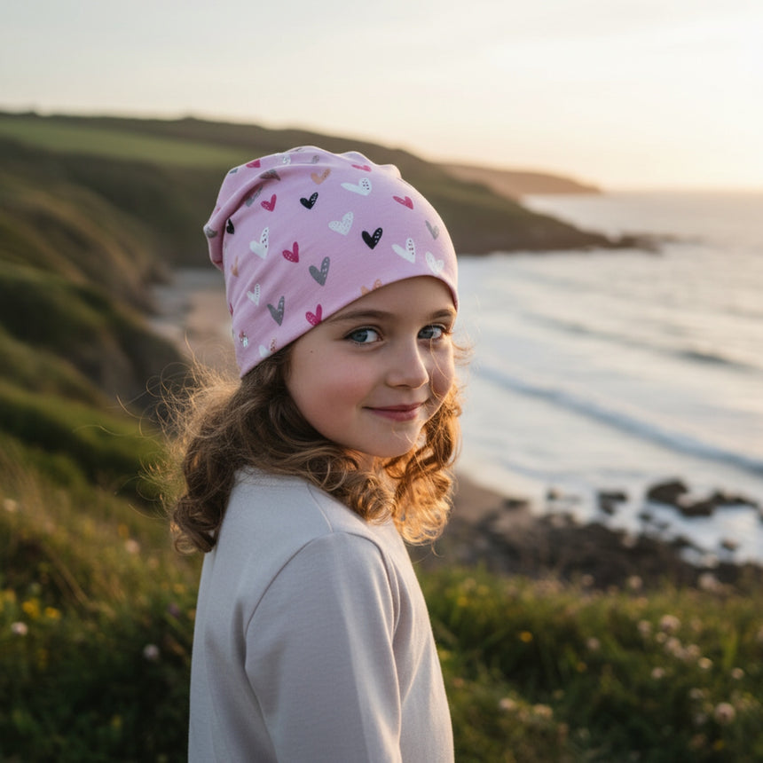 Mädchen trägt Wollhuhn Öko Long Beanie / Wendemütze in Rosa mit Herzen, steht auf Küstenklippe bei Sonnenuntergang, umgeben von Meer und Hügeln.
