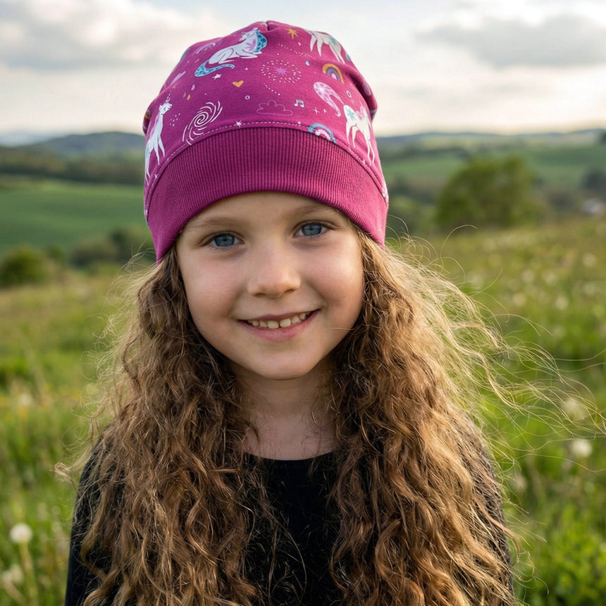 Mädchen mit blauer Augen trägt die Wollhuhn Dünne Öko Beanie-Mütze, pink mit Einhornmotiv, in einem grünen Feld. Handgefertigt, dehnbar, ideal für Kinder.