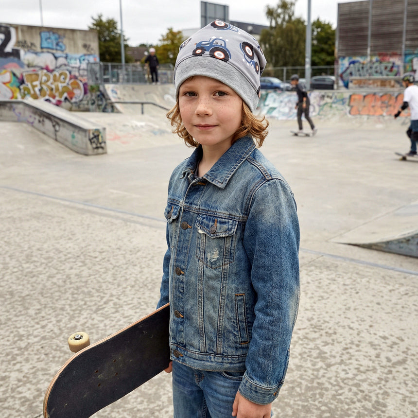 Kind im Skatepark trägt Wollhuhn Dünne Öko Beanie-Mütze Sommer/Übergang Einlagig Maxi Traktor/Trecker Grau, hält Skateboard, Graffiti im Hintergrund.