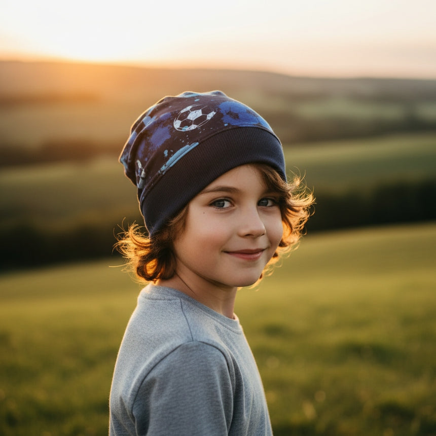 Kind mit Wollhuhn Dünne Öko Beanie-Mütze, einlagig und mit Fußballmuster, steht in einem Sonnenuntergangsfeld. Ideal für Übergang, handgefertigt, bequem und dehnbar.
