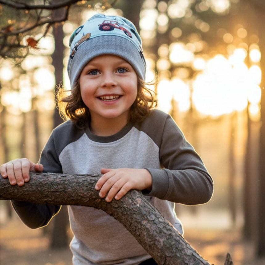 Kind mit Wollhuhn Dünne Öko Beanie-Mütze Maxi Trecker, lächelt auf Baumstamm, von Sonnenlicht umgeben. Handgemachte, bequeme Mütze, ideal für Übergangszeit.