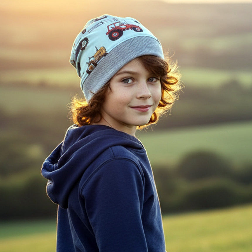 Kind trägt Wollhuhn Dünne Öko Beanie-Mütze mit Traktormuster in hellblau, lächelt im Freien, umgeben von grünen Feldern bei goldenem Licht.